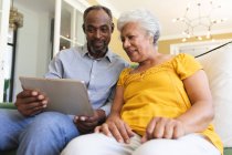 Primer plano de una feliz pareja de ancianos afroamericanos jubilados en casa sentados en un sofá en su sala de estar, utilizando una tableta juntos y sonriendo, pareja aislante durante coronavirus covid19 pandemia - foto de stock