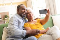 Счастливая пожилая афроамериканская пара в своей гостиной, сидящая на диване, женщина, держащая смартфон, оба смотрят на телефон вместе, делают селфи и улыбаются, пара изолируется во время пандемии coronavirus covid19 — стоковое фото