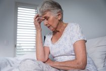 Закриття старої кавказької жінки вдома сидячи в ліжку з головним болем, тримаючи голову закритими очима, самоізолюючись під час коронавірусної кочівлі ковідій-19 пандемії. — стокове фото