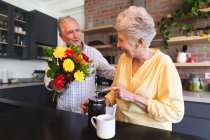 Старша кавказька пара, яка відпочиває на пенсії, стоїть на кухні в сонячний день, жінка готує каву і чоловік дарує їй букет квітів вдома, ізолюючись під час коронавірусу ковідій ковідемії. — стокове фото