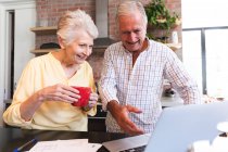 Una pareja de ancianos jubilados caucásicos en casa de pie en una mesa en su cocina, hablando y sonriendo, utilizando un ordenador portátil juntos, pareja aislante durante coronavirus covid19 pandemia - foto de stock