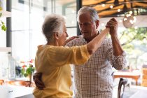 Счастливая пожилая белая пара дома, держащаяся за руки, танцующая вместе на кухне и улыбающаяся, дома вместе изолирующая во время пандемии коронавируса — стоковое фото