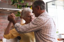 Primer plano de la feliz pareja de ancianos caucásicos jubilados en casa tomados de la mano, bailando juntos en su cocina y sonriendo, en casa juntos aislando durante coronavirus covid19 pandemia - foto de stock