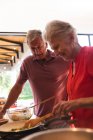 Primo piano di una coppia caucasica anziana in pensione felice a casa, preparare il cibo e sorridere nella loro cucina, la donna che cucina le verdure in una padella, l'uomo appoggiato, guardare e parlare ed entrambi sorridenti. — Foto stock