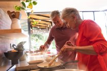 Feliz pareja caucásica jubilada en casa, preparando comida y sonriendo en su cocina, la mujer cocinando verduras en una sartén, el hombre apoyado, mirando y hablando y ambos sonriendo - foto de stock