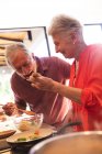 Счастливая пожилая кавказская пара дома, готовящая еду на своей кухне вместе, женщина, дающая мужчине полный рот еды из деревянной ложки, дома вместе изолирующая во время пандемии коронавируса — стоковое фото