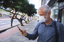 Старший кавказский мужчина гуляет по улицам города в течение дня, носит маску против коронавируса, ковид 19 и использует смартфон. — стоковое фото