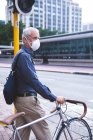 Hombre caucásico mayor por las calles de la ciudad durante el día, usando una máscara facial contra el coronavirus, covid 19, moviendo su bicicleta. - foto de stock