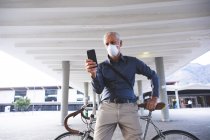 Старший кавказский мужчина днем ходит по улицам города в маске против коронавируса, ковид 19, сидит на велосипеде и пользуется смартфоном. — стоковое фото