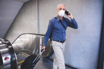 Senior Caucasian man, wearing a face mask against coronavirus, covid 19, using an escalator in a metro station, talking on his smartphone, and pulling a suitcase. — Stock Photo