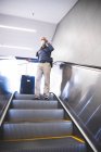 Старший кавказький чоловік, одягнений в маску обличчя проти коронавірусу, коїд 19, використовуючи ескалатор на станції метро, розмовляючи на своєму смартфоні, і тягнучи валізу.. — стокове фото