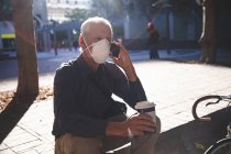 Älterer kaukasischer Mann, der tagsüber auf den Straßen der Stadt unterwegs ist, Gesichtsmaske gegen Coronavirus trägt, covid 19, auf der Treppe sitzt, eine Tasse Kaffee zum Mitnehmen in der Hand hält und sein Smartphone benutzt. — Stockfoto