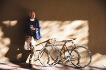 Uomo anziano caucasico in giro per le strade della città durante il giorno, indossando una maschera contro il coronavirus, covid 19, appoggiato al muro e tenendo una tazza di caffè da asporto mentre la sua bicicletta è in piedi accanto a lui. — Foto stock
