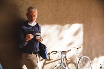 Älterer kaukasischer Mann, der tagsüber auf den Straßen der Stadt unterwegs ist, Gesichtsmaske gegen Coronavirus trägt, 19 Jahre alt, an die Wand gelehnt, eine Tasse Kaffee zum Mitnehmen hält und sein Smartphone benutzt. — Stockfoto
