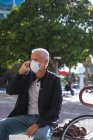 Homme caucasien âgé dehors et autour dans les rues de la ville pendant la journée, portant un masque facial contre le coronavirus, covid 19, assis sur un banc et en utilisant son smartphone. — Photo de stock
