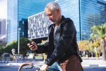 Homem caucasiano sênior nas ruas da cidade durante o dia, sentado em sua bicicleta e usando um smartphone . — Fotografia de Stock