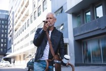 Homem caucasiano sênior fora e sobre nas ruas da cidade durante o dia, sentado em sua bicicleta e usando um smartphone, segurando uma xícara de café takeaway . — Fotografia de Stock