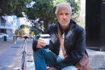 Senior Caucasian man, wearing casual clothes, out and about in the city streets during the day, sitting on stairs, holding a cup of takeaway coffee and looking straight into a camera. — Stock Photo