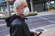 Homem caucasiano sênior nas ruas da cidade durante o dia, usando uma máscara facial contra coronavírus, vívido 19, usando um smartphone . — Fotografia de Stock