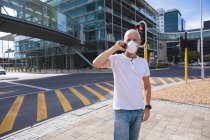 Uomo anziano caucasico in giro per le strade della città durante il giorno, indossando una maschera contro il coronavirus, covid 19, utilizzando uno smartphone. — Foto stock