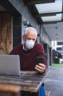 Älterer kaukasischer Mann sitzt an einem Tisch auf einer Kaffeeterrasse und trägt eine Gesichtsmaske gegen Coronavirus, Covid 19, mit Smartphone und Laptop. — Stockfoto