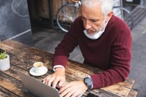 Senior Caucasian man sitting at a table at a coffee terrace, wearing a face mask against coronavirus, covid 19, using a laptop computer. — Stock Photo