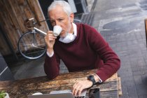Senior Caucasian man sitting at a table at a coffee terrace, wearing a face mask against coronavirus, covid 19, drinking coffee and using a laptop computer. — Stock Photo