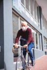 Homem caucasiano sênior nas ruas da cidade durante o dia, usando uma máscara facial contra o coronavírus, vívido 19, andando de bicicleta . — Fotografia de Stock