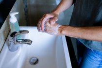 Средняя часть женщины дома в ванной комнате в дневное время моет руки в раковине с помощью мыла, бутылка с жидким мылом рядом с ней, защита от коронавируса Covid-19 инфекции и пандемии. — стоковое фото