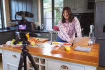 Vlogger mujer caucásica en casa en su cocina, demostrando la preparación de recetas de alimentos delante de una cámara para su blog en línea. Distanciamiento social y autoaislamiento en cuarentena. - foto de stock