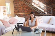 Vlogger mujer caucásica en casa, en su sala de estar utilizando un ordenador portátil para preparar su blog en línea. Distanciamiento social y autoaislamiento en cuarentena. - foto de stock