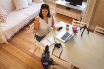 Mulher branca vlogger em casa, em sua sala de estar usando uma câmera e um smartphone, um laptop e uma câmera para preparar seu blog online. Distanciamento social e auto-isolamento em quarentena . — Fotografia de Stock