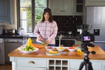 Femme vlogger caucasienne à la maison dans sa cuisine, démontrant la préparation de recettes alimentaires devant une caméra pour son blog en ligne. Distance sociale et isolement personnel en quarantaine. — Photo de stock