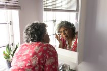 Une femme afro-américaine âgée passe du temps à la maison, à se distancer socialement et à s'isoler en quarantaine pendant l'épidémie de coronavirus covid 19, se regardant dans un miroir et touchant son visage — Photo de stock