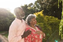 Um casal afro-americano sênior que passa tempo em seu jardim juntos, distanciamento social e auto-isolamento em quarentena durante coronavírus covid 19 epidemia, abraçando e olhando para longe — Fotografia de Stock