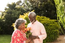 Um casal afro-americano sênior que passa um tempo em seu jardim juntos, distanciamento social e auto-isolamento em quarentena durante a epidemia do coronavírus covid 19, o homem segurando um buquê de flores, sorrindo e dando-os à mulher — Fotografia de Stock
