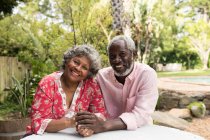 Porträt eines älteren afroamerikanischen Paares, das während der Coronavirus-Epidemie von 19 Jahren Zeit in seinem Garten verbringt, soziale Distanzierung und Selbstisolierung in Quarantäne, Händchenhalten, in die Kamera schauen und lächeln — Stockfoto