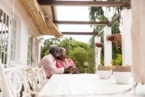 Старшая афроамериканская пара проводит время в своем саду вместе, социальное дистанцирование и самоизоляция в карантинной изоляции во время эпидемии коронавируса, улыбаясь и отводя взгляд, с бокалами красного вина на столе — стоковое фото
