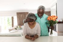 Un couple afro-américain sénior passe du temps à la maison ensemble, distance sociale et isolement personnel en quarantaine pendant l'épidémie de coronavirus covid 19, l'homme tenant un bouquet de fleurs, la femme tenant un smartphone — Photo de stock