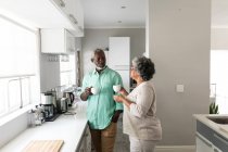 Старшая афроамериканская пара, проводящая время дома вместе, социальное дистанцирование и самоизоляция в карантинной изоляции во время эпидемии коронавируса, стоящая на кухне, разговаривающая, держащая чашки — стоковое фото