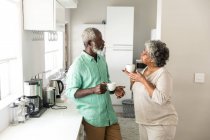 Un couple afro-américain âgé passe du temps à la maison ensemble, la distance sociale et l'isolement personnel en quarantaine pendant l'épidémie de coronavirus covid 19, debout dans la cuisine, parlant, tenant des tasses — Photo de stock