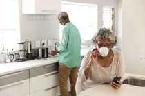 Ein älteres afroamerikanisches Paar, das während der Coronavirus-Epidemie 19 Zeit zu Hause verbringt, soziale Distanzierung und Selbstisolierung in Quarantäne, die Frau benutzt ein Smartphone und trinkt Kaffee — Stockfoto