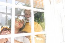 Старшая афроамериканская пара проводит время дома вместе, социальное дистанцирование и самоизоляция в карантинной изоляции во время эпидемии коронавируса ковида 19, глядя в окно улыбаясь, обнимаясь — стоковое фото