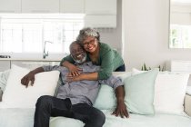 Retrato de una pareja afroamericana mayor pasando tiempo en casa juntos, distanciamiento social y aislamiento en cuarentena durante la epidemia de coronavirus covid 19, mirando a la cámara, sonriendo, abrazando - foto de stock