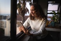 Caucasian woman spending time at home self isolating and social distancing in quarantine lockdown during coronavirus covid 19 epidemic, sitting by the window and smiling while using tablet. — Stock Photo