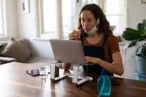 Caucasian woman spending time at home, wearing a face mask against coronavirus, covid 19, sitting by her desk and working, using her laptop and earphones. Social distancing and self isolation in quarantine lockdown. — Stock Photo