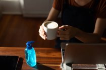Mid section of woman spending time at home, sitting by her desk, taking a break from work and holding a cup of coffee after cleaning her hands with a sanitizer. Social distancing and self isolation in quarantine lockdown. — Stock Photo