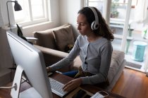 Mulher caucasiana passando tempo em casa, usando fones de ouvido, sentada ao lado de sua mesa e trabalhando usando seu computador. Distanciamento social e auto-isolamento em quarentena . — Fotografia de Stock