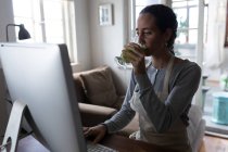 Кавказька жінка проводить час удома, сидячи за своїм столом і працюючи за комп'ютером, п'ючи коктейль. Соціальна дистанція і самоізоляція в карантинному блокуванні.. — стокове фото