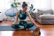 Caucasian woman spending time at home, wearing sportswear, sitting on a yoga mat and stretching up, joining online yoga course, using her tablet. Social distancing and self isolation in quarantine lockdown. — Stock Photo